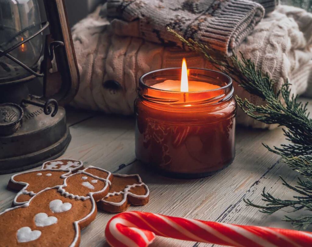 Holiday scented candle in amber colored mason jar on table in front of folded sweater and mittens next to gingerbread cookies, a candy cane and a lantern.