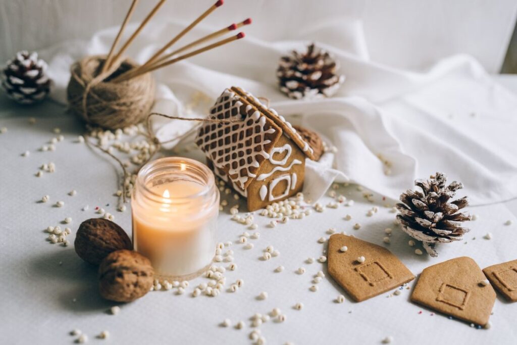 Holiday scented candle in clear mason jar on table with gingerbread house and pinecones against white background.