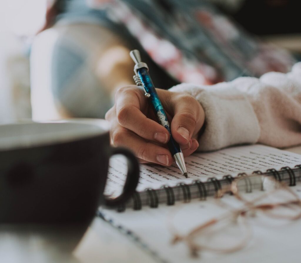 Someone's hand writing in a journal notebook with eyeglasses to the side and a coffee mug in the front.