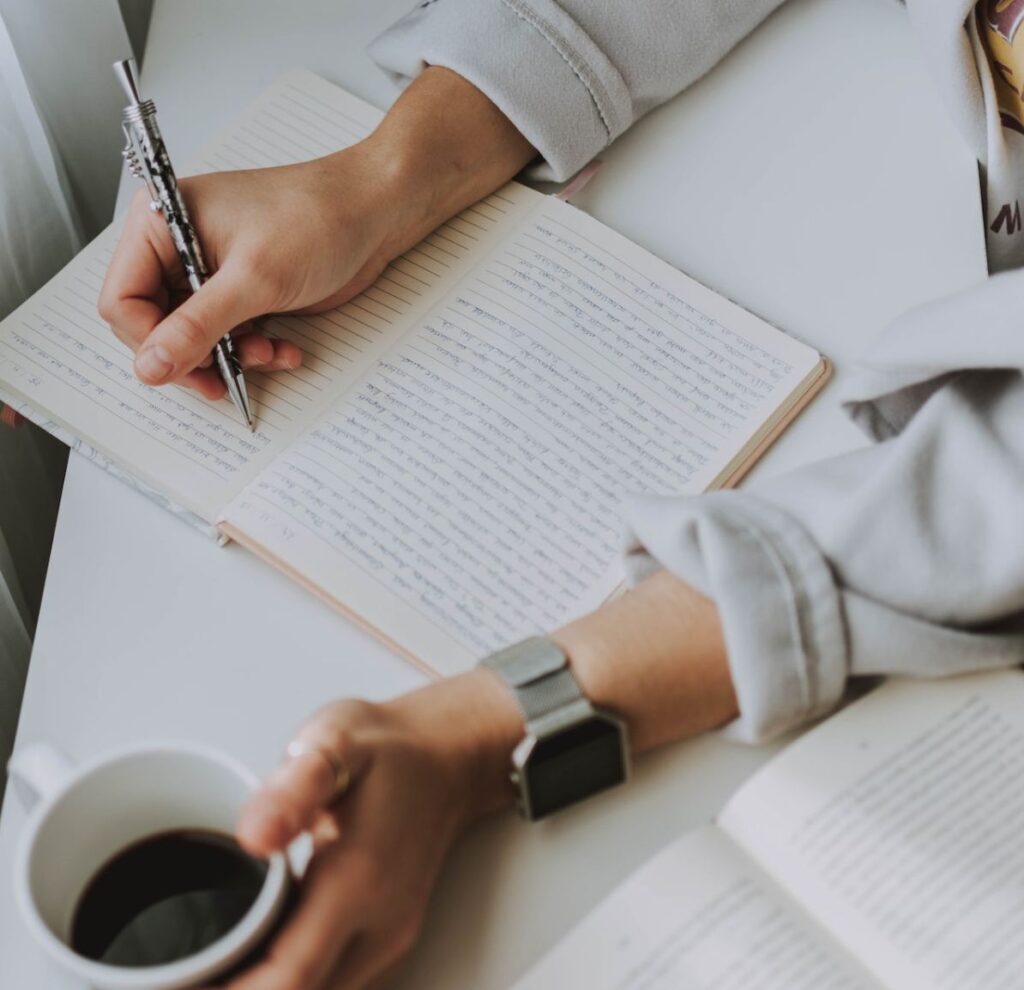 Someone's hand writing in a journal notebook with a cup of coffee in the other hand. 